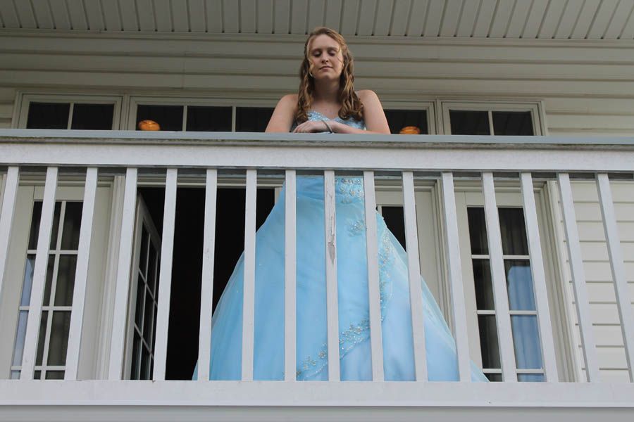Woman in a light blue formal dress leans on a white balcony railing.