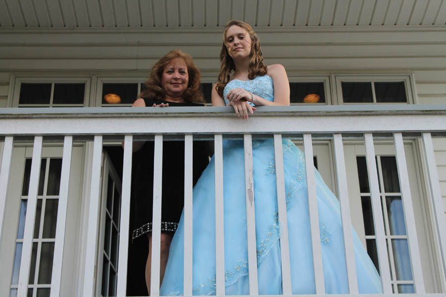 Woman in blue gown stands with another woman on a white porch, leaning on the railing.
