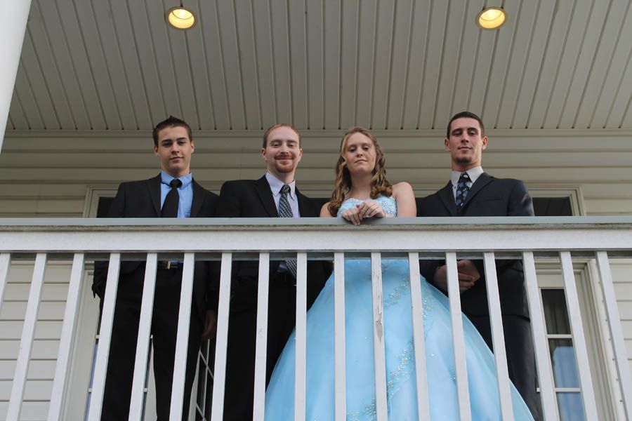 Four people on a white porch: three men in suits flanking a woman in a blue gown.