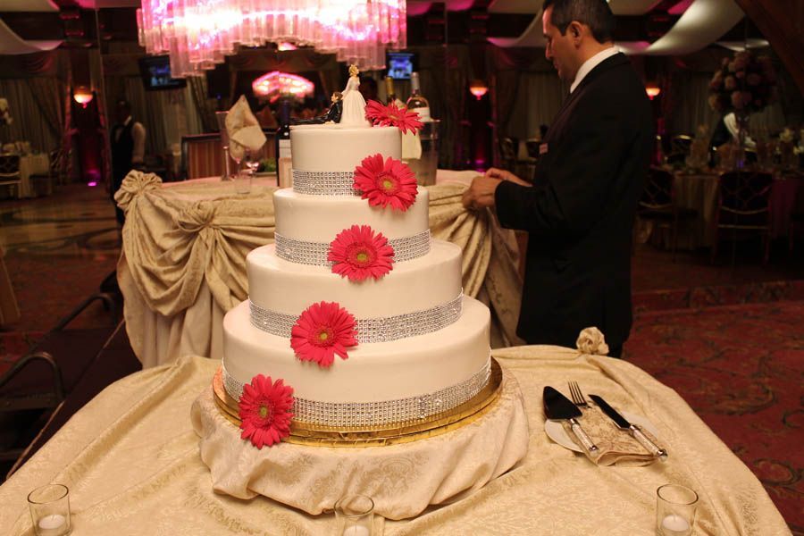 Four-tiered white wedding cake with pink flowers and silver trim on a table, a man in a suit stands next to it.