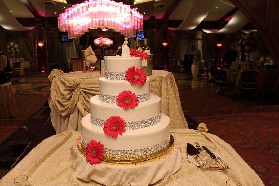 Four-tiered wedding cake with pink flowers and silver trim on a table in a ballroom.