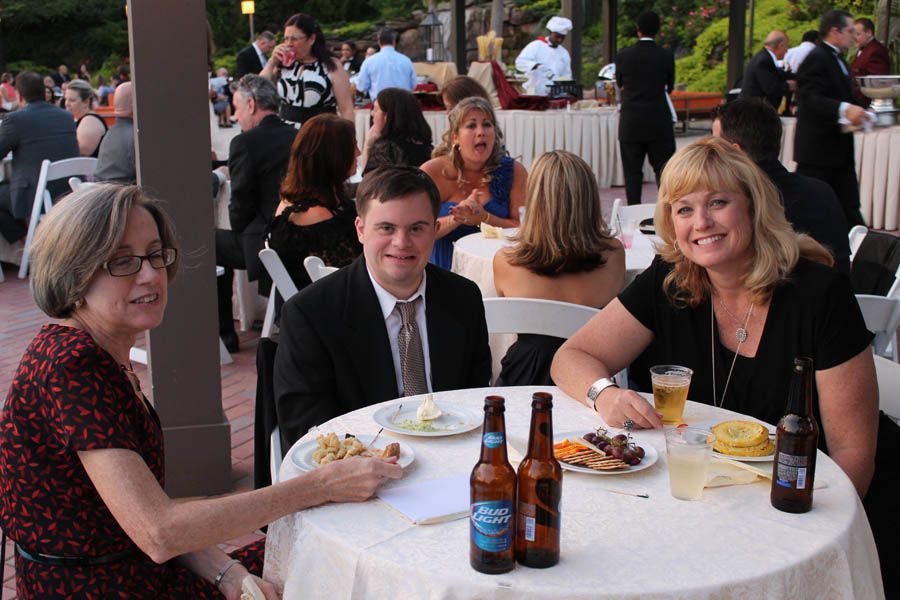 Three people at a table; man with Down syndrome smiles between two women, outdoor party.