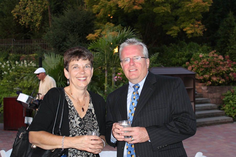 Woman and man smiling, holding drinks, outdoors near greenery. Man in suit, woman in patterned top.