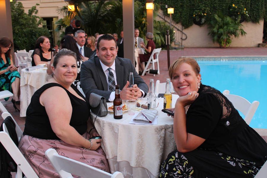 Three people at a round table near a pool: two women and one man, smiling, at an outdoor event.
