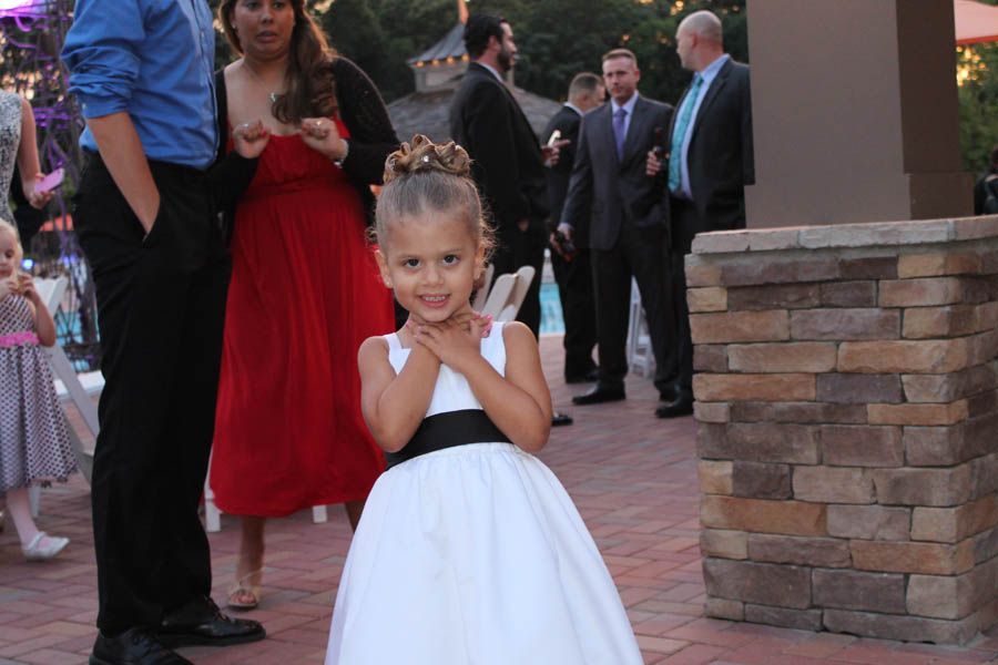 Smiling girl in white dress with black sash; hands on chest, posing at event. People in background.