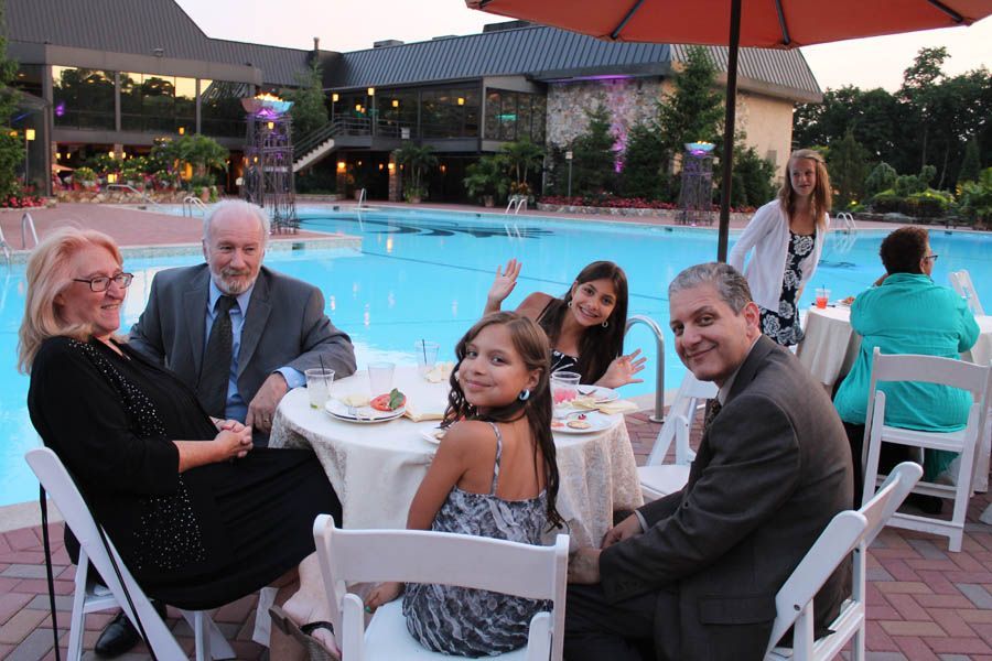 People at outdoor table near pool, waving. Building and umbrella in background.