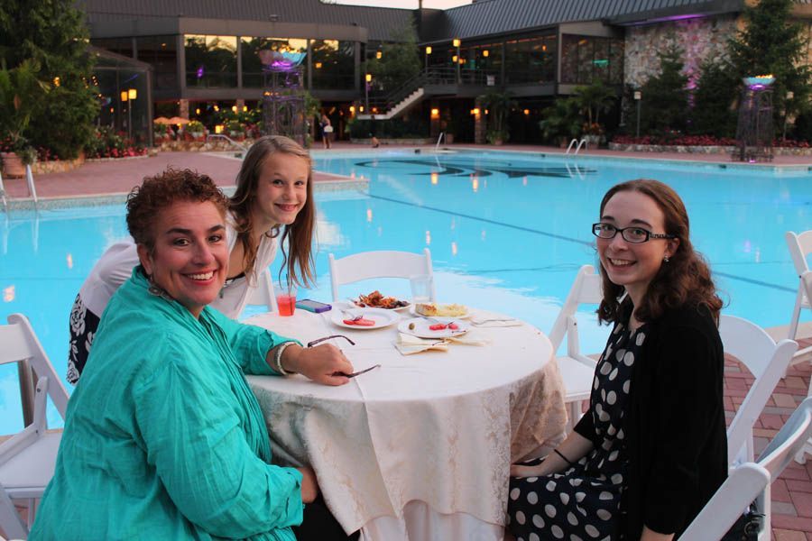 Three people at a table by a pool; one woman in teal smiles, another girl and woman smile.