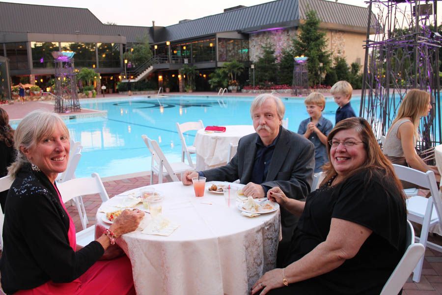 Three people sit at a round table by a pool, smiling. Evening outdoor setting, food and drinks.