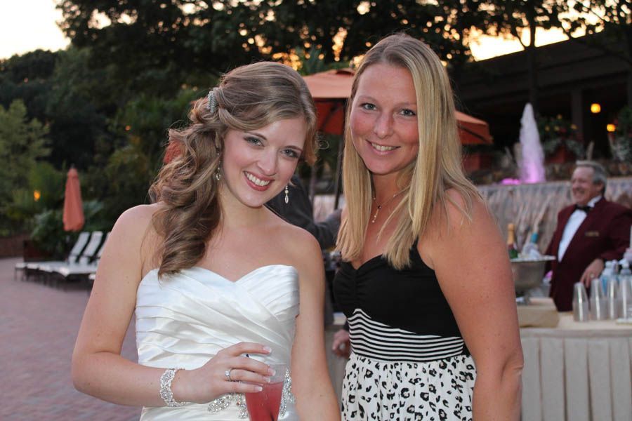 Bride in white dress smiles next to woman in black and white dress at outdoor event with fountain.