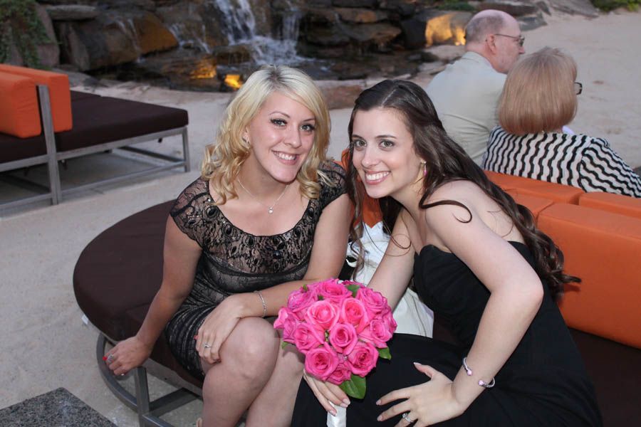Two women with smiles pose, one holds pink roses. Outdoor setting, waterfall backdrop.