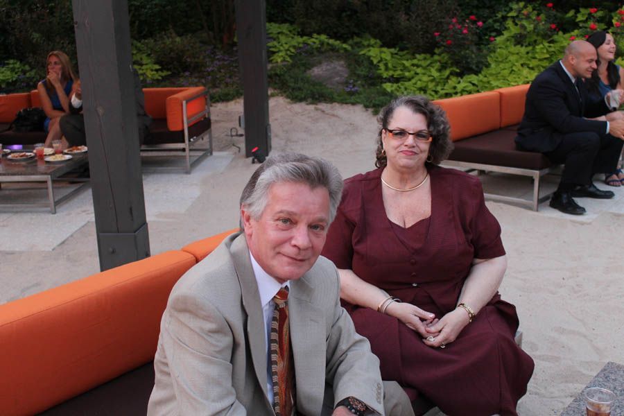 Man in tan suit and woman in burgundy dress seated outdoors, smiling. Orange benches in background.