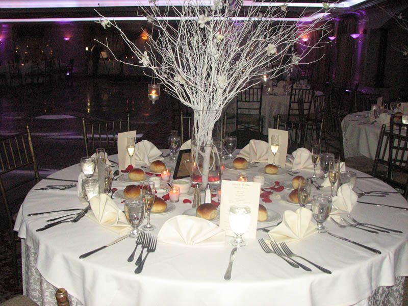 Round table set for a formal event, featuring white tree centerpiece, place settings with folded napkins, and soft lighting.