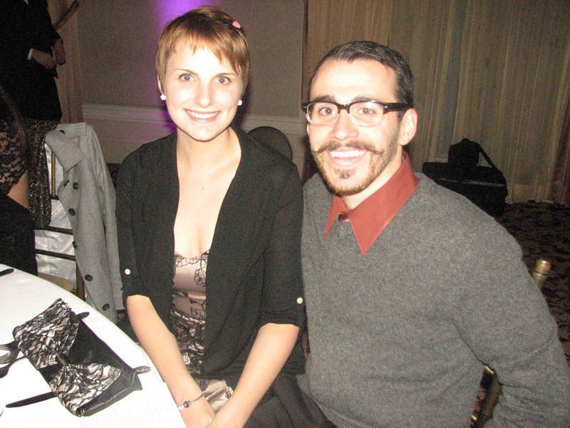 Woman and man smiling at a table. Woman has short hair and a black sweater. Man has glasses and a mustache.