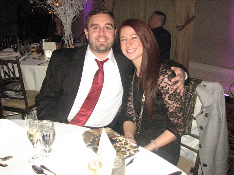 Man in black suit and red tie, arm around woman in black lace dress, smiling at a formal event.
