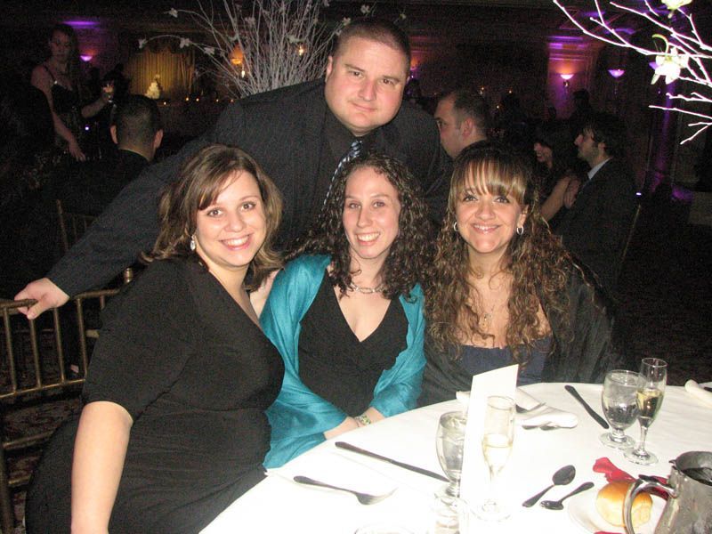 Four people at a table, smiling. A man stands behind them. Festive setting with white floral decor.