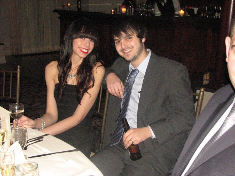 Woman in black dress and man in suit at a table, smiling. Holding drinks, lit room, formal event.