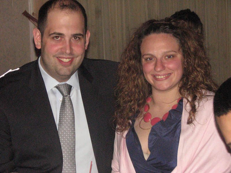 Smiling couple posing together indoors; man in suit, woman in blue dress and pink shawl.