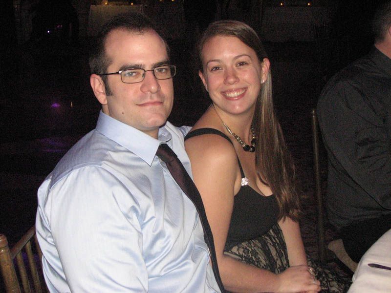 Man in blue shirt and woman in black dress sit together, smiling.
