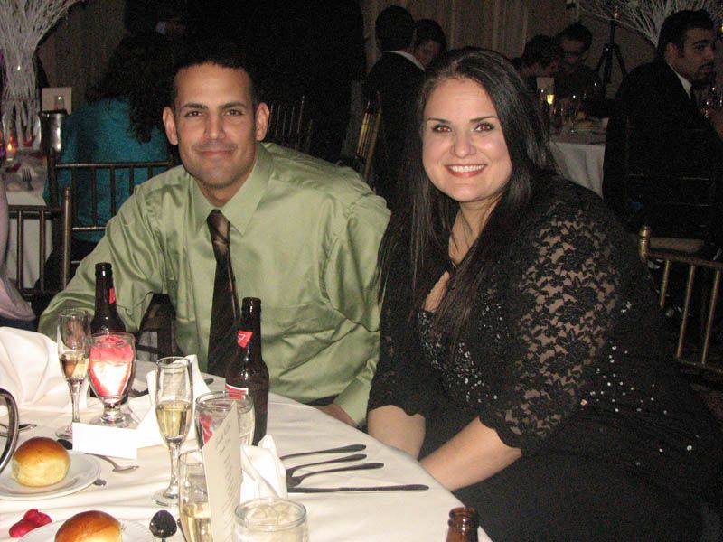 Man and woman seated at a formal table, smiling. Man in green shirt, woman in black lace. Tables with food and drinks.