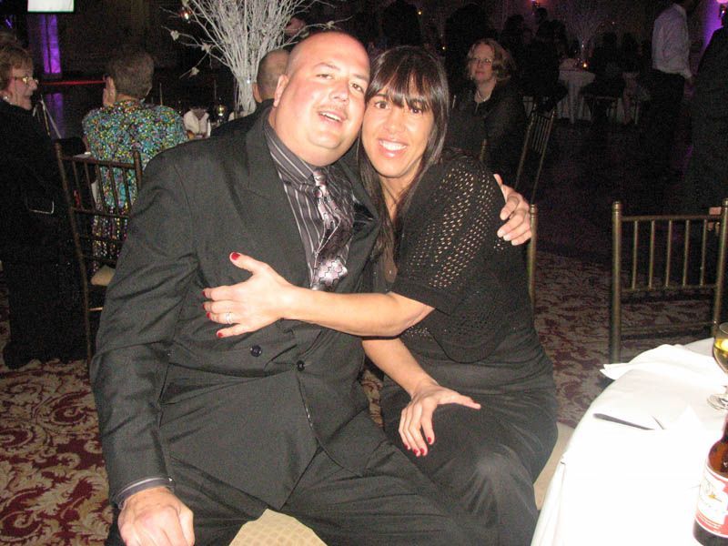 Man and woman embrace at a formal event, smiling. Man wears a suit, woman in black top and pants.