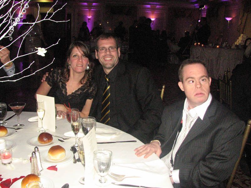 People seated at a decorated table at an event. Man in suit smiles, another man and woman look at camera.