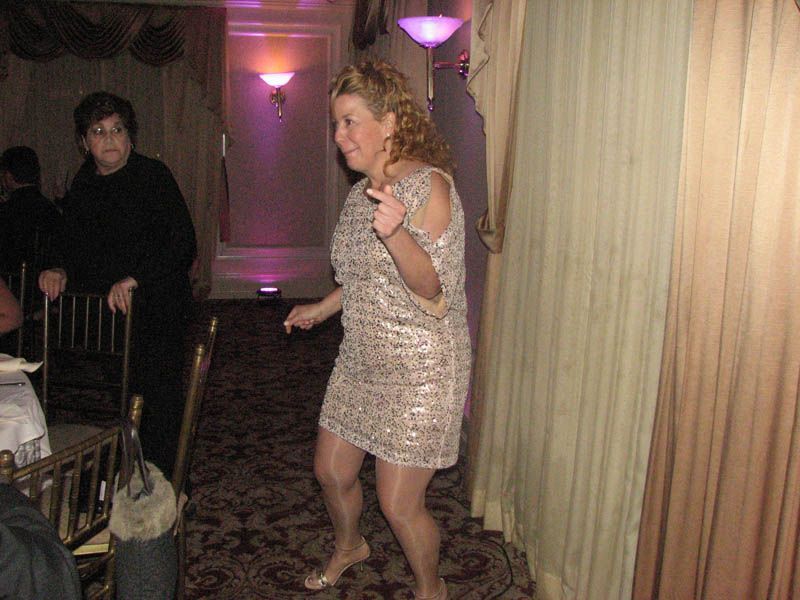 Woman in sequined dress dancing near curtain; another person observes nearby.