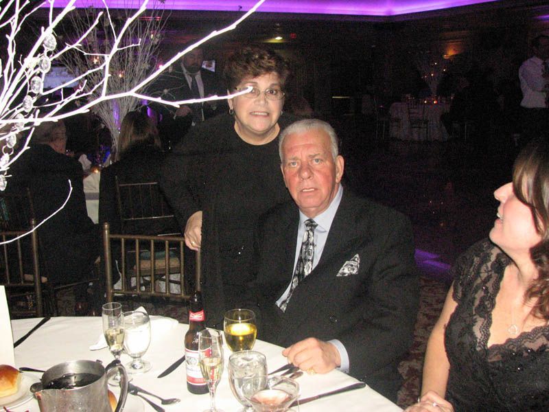 Couple at a formal event; woman standing behind man seated at a decorated table, smiling.