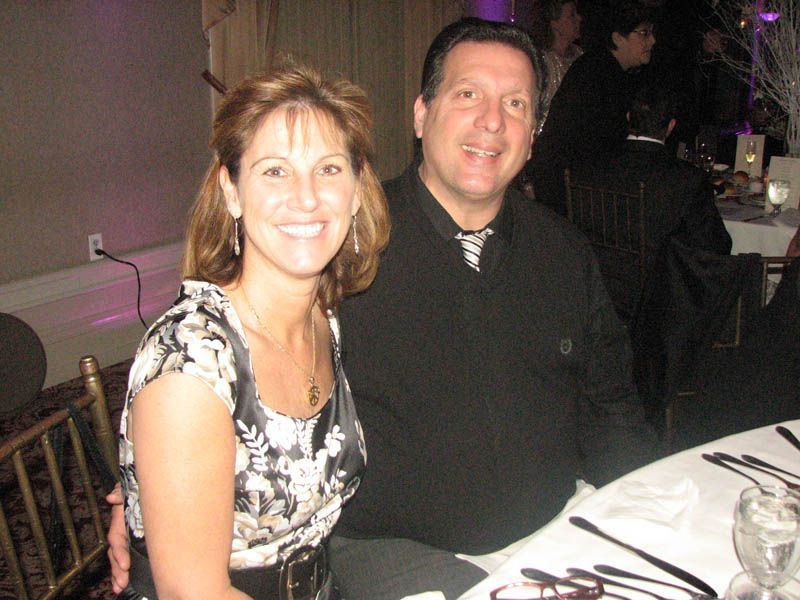 Smiling couple at a formal event; woman in floral top, man in black shirt, sitting at a table.
