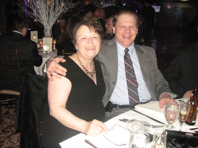 Couple smiling at a formal event, woman in black dress, man in gray suit, arms around each other at a table.
