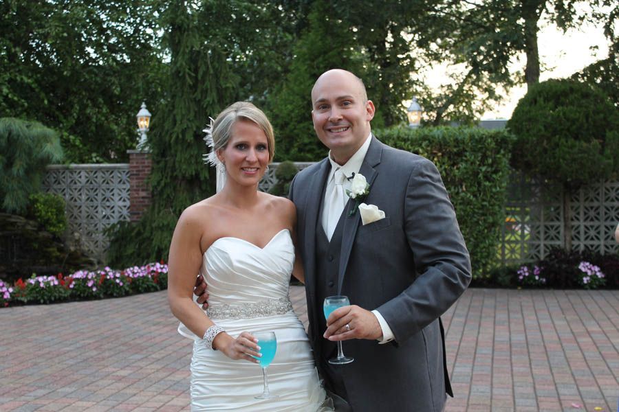 Bride and groom smiling, holding drinks, in wedding attire, outdoors on a brick patio.