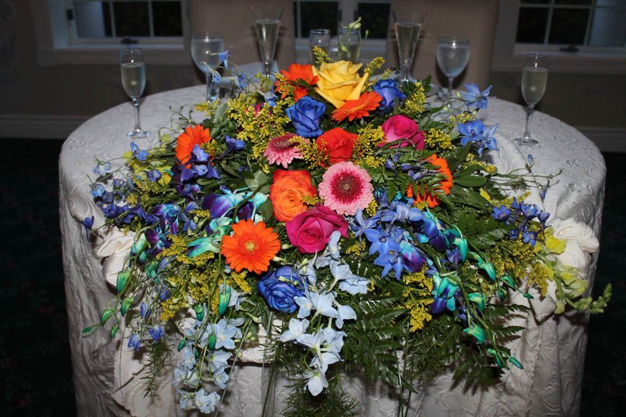 Floral centerpiece on a round, draped table with champagne flutes; flowers include orange, blue, yellow, and pink blooms.