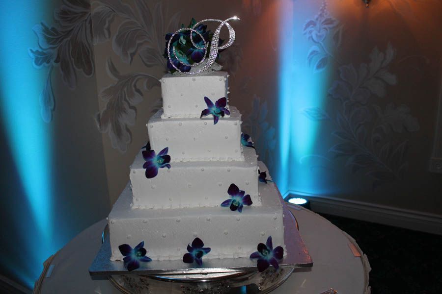 Four-tiered white wedding cake with purple flowers and a monogram topper on a table with blue lighting.