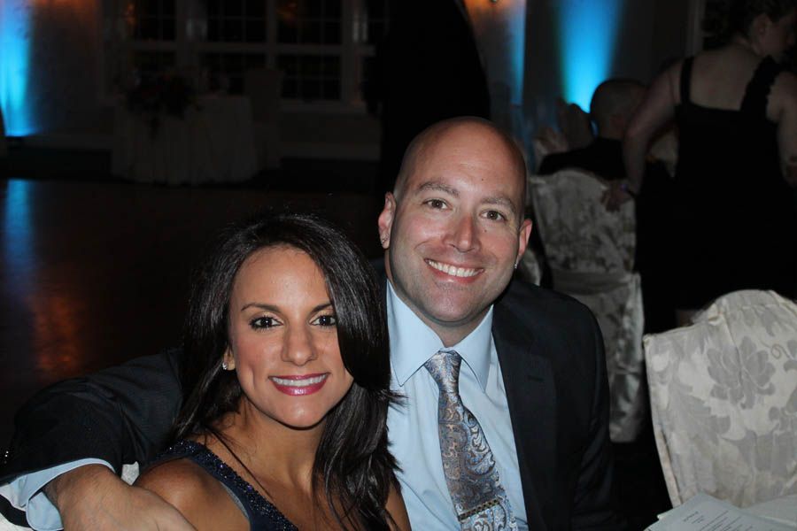 Couple smiling at camera; man in suit and tie, woman in dark top, dim interior, soft lighting.