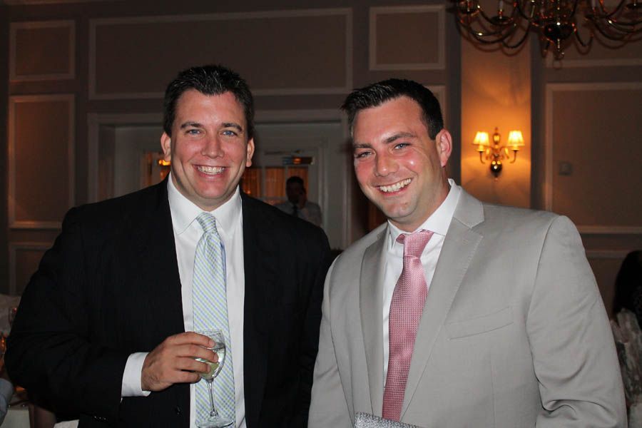 Two men smiling in suits, light blue and pink ties, indoors with chandelier.