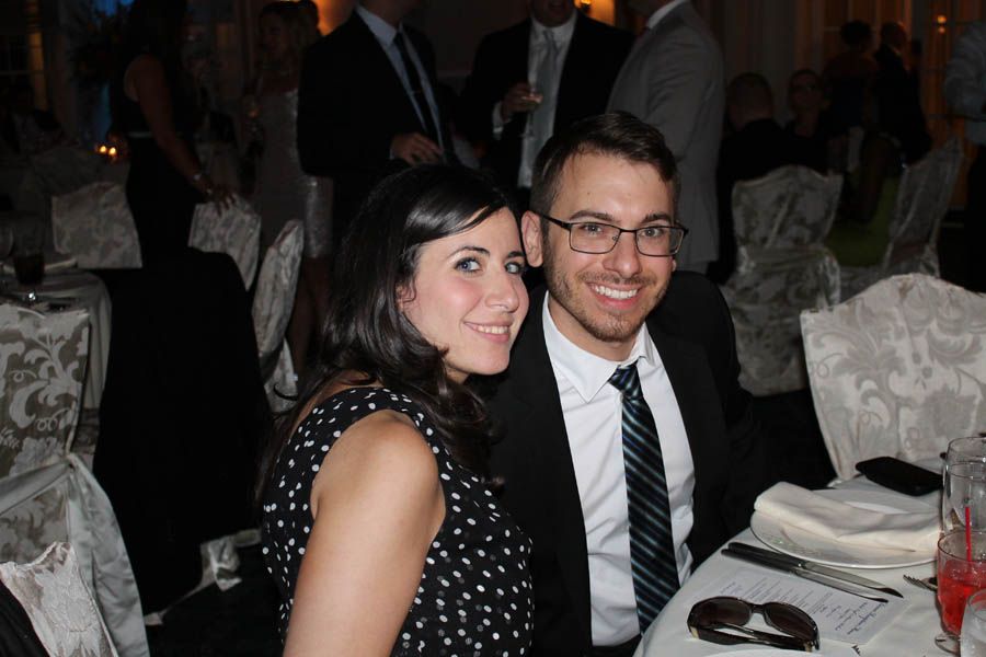 Smiling couple at a formal event; woman in polka dots, man in a suit. Tables, guests in background.