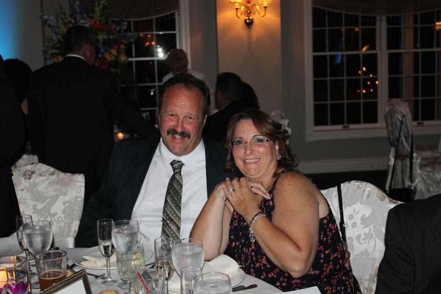 Couple at a table, smiling at the camera. Man in suit jacket, tie. Woman in patterned dress. Formal event.