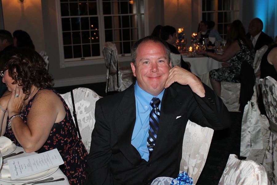 Man in suit at a formal event, seated, smiling with hand on his chin, light blue shirt, patterned tie.