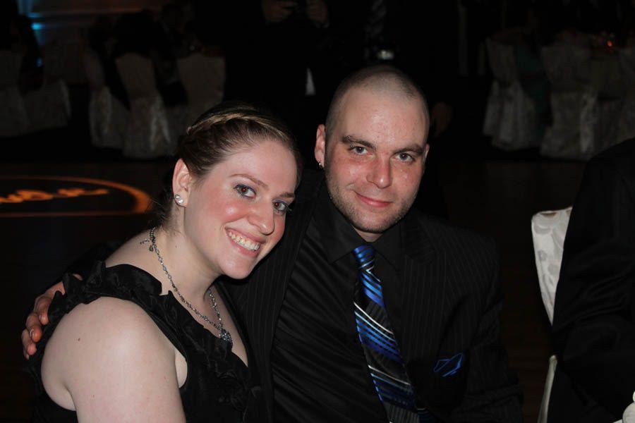 Woman in a black dress smiles next to a man in a black suit with a patterned tie. They are seated indoors.