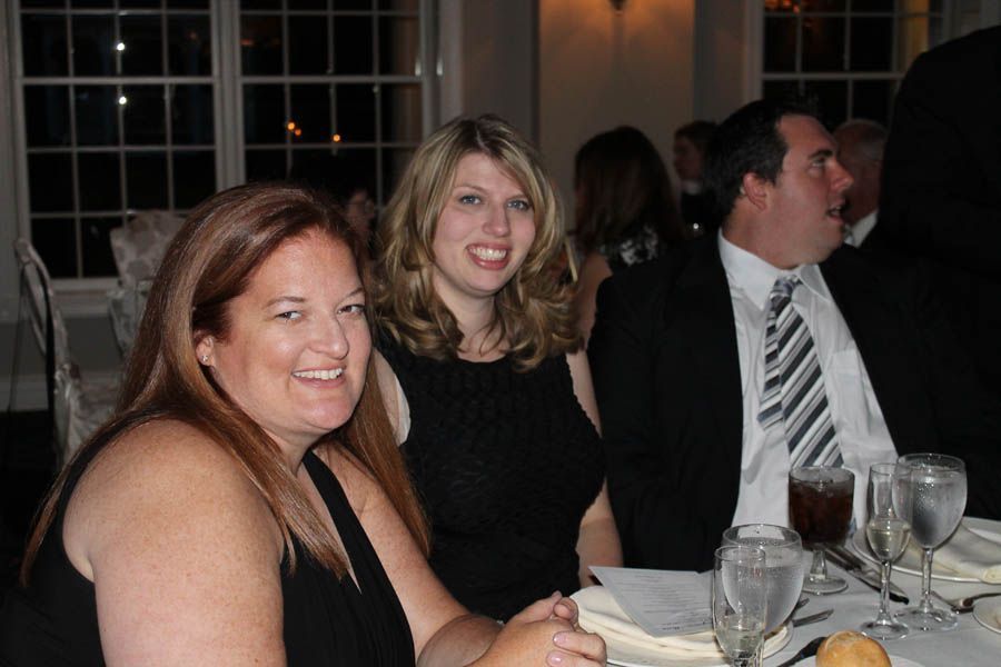 Three people at a formal dinner table, smiling. Two women and a man. White tablecloth, glasses. Dim lighting.