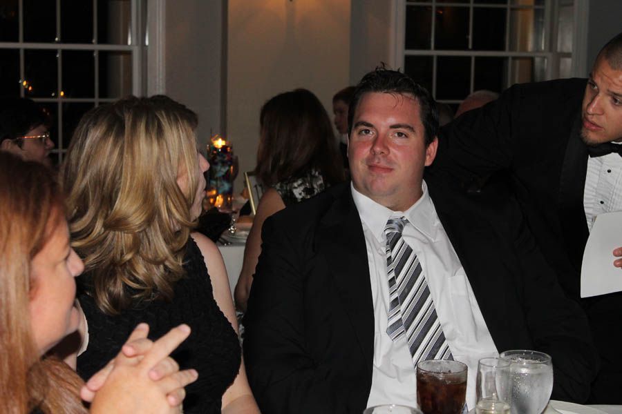 Man in a suit smiles at a table; others seated. Formal setting with window.
