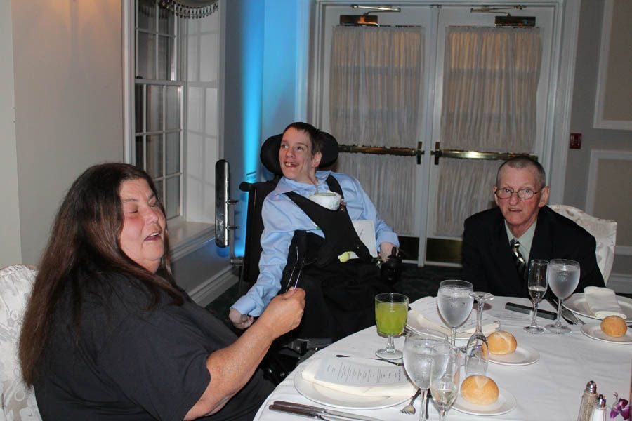 Three people at a banquet table: Woman laughs, man in a wheelchair smiles, and older man looks on.