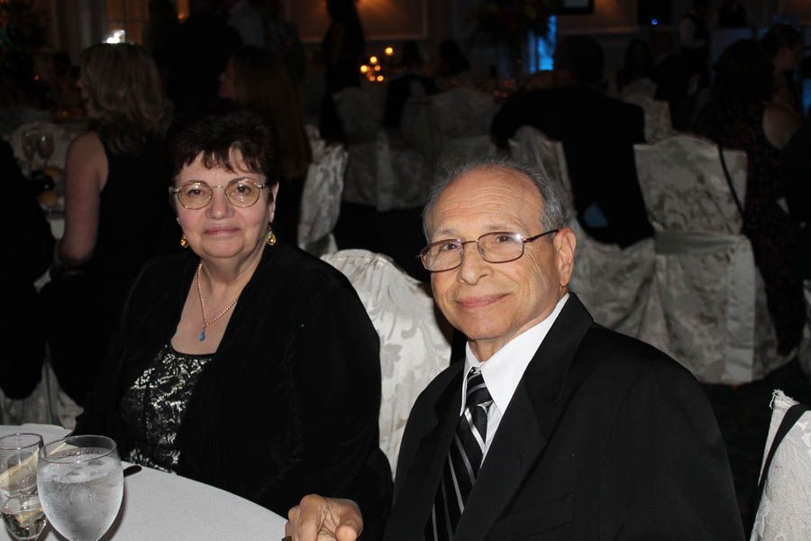 Older couple at a formal event, seated at a table. Woman in black top, man in suit, both smiling.