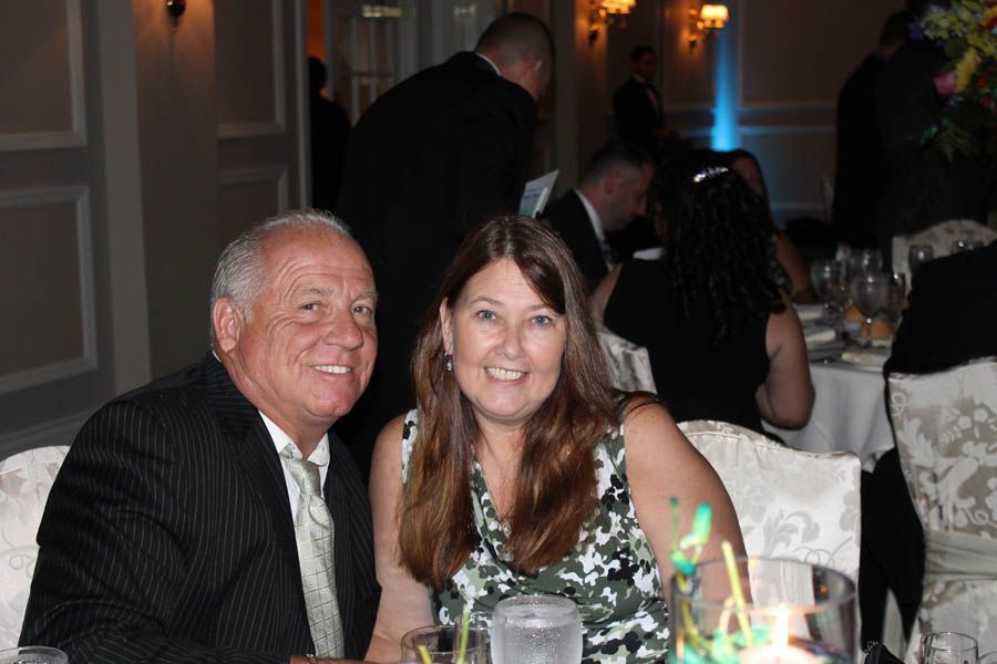 Man and woman smiling at a table, likely a formal event. She is in a floral top; he is in a suit.
