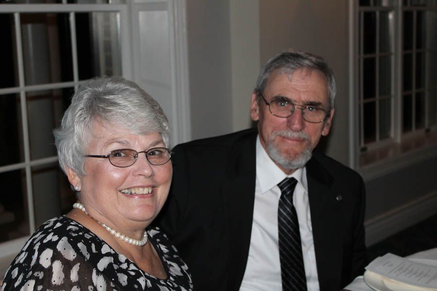 Couple smiling; woman in patterned top, man in suit, glasses. Indoor setting, near a window.