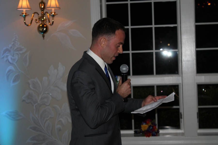 Man in a suit speaks into a microphone, reading from papers, near a window at an event.