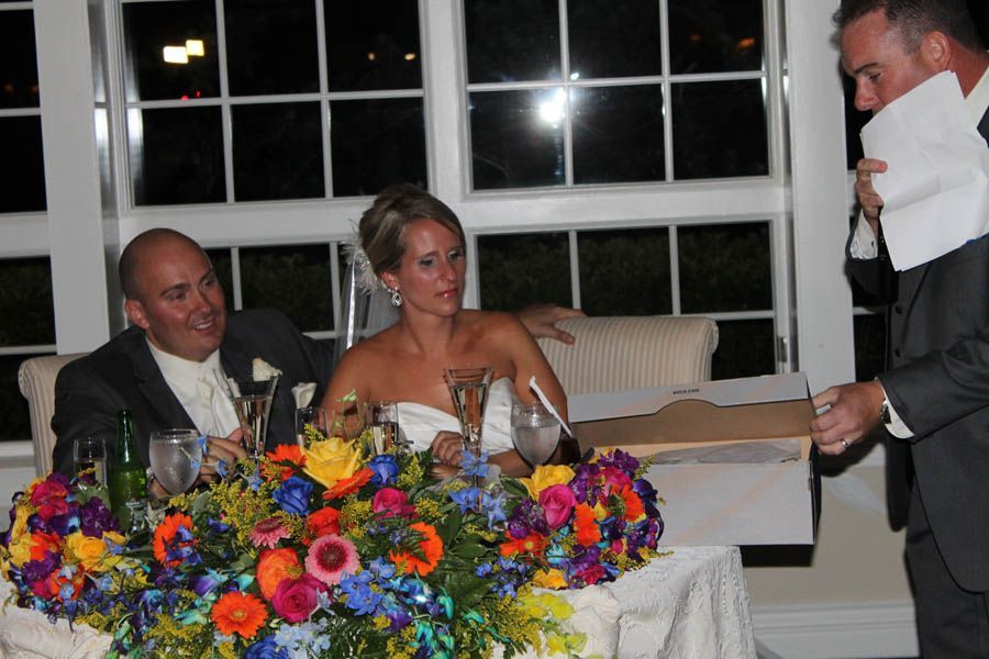 Bride and groom at a wedding reception table; man gives a speech, colorful flowers.