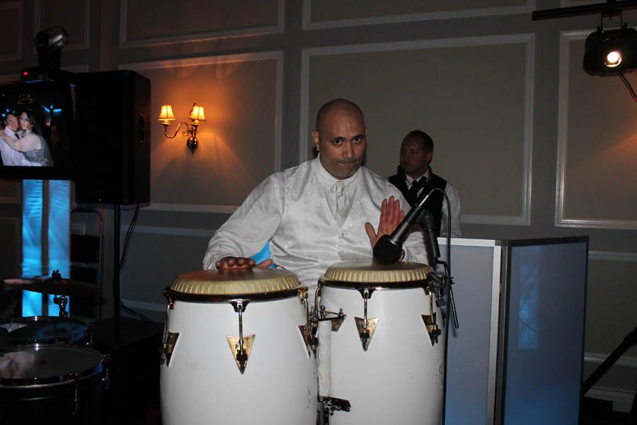 Man playing conga drums at an event, wearing a white shirt and vest. Black microphone, neutral expression.