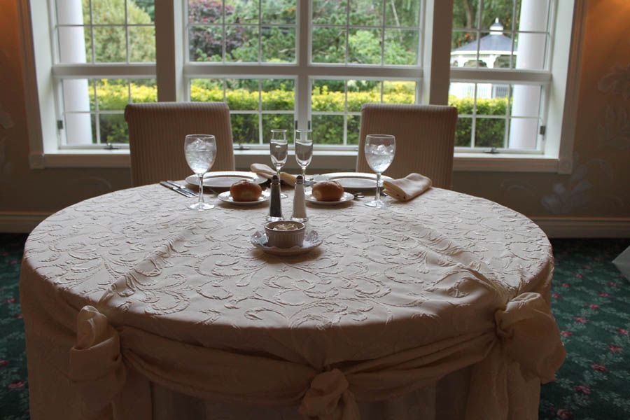Round table set for a meal, draped in lace, with window view of garden.