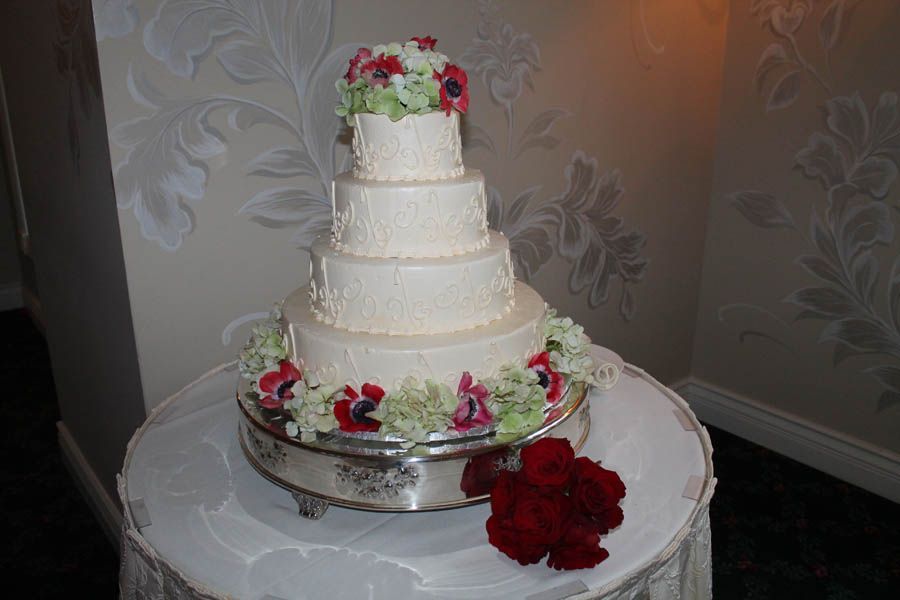 Four-tiered wedding cake with floral decorations on a silver stand, set against a floral wallpaper.