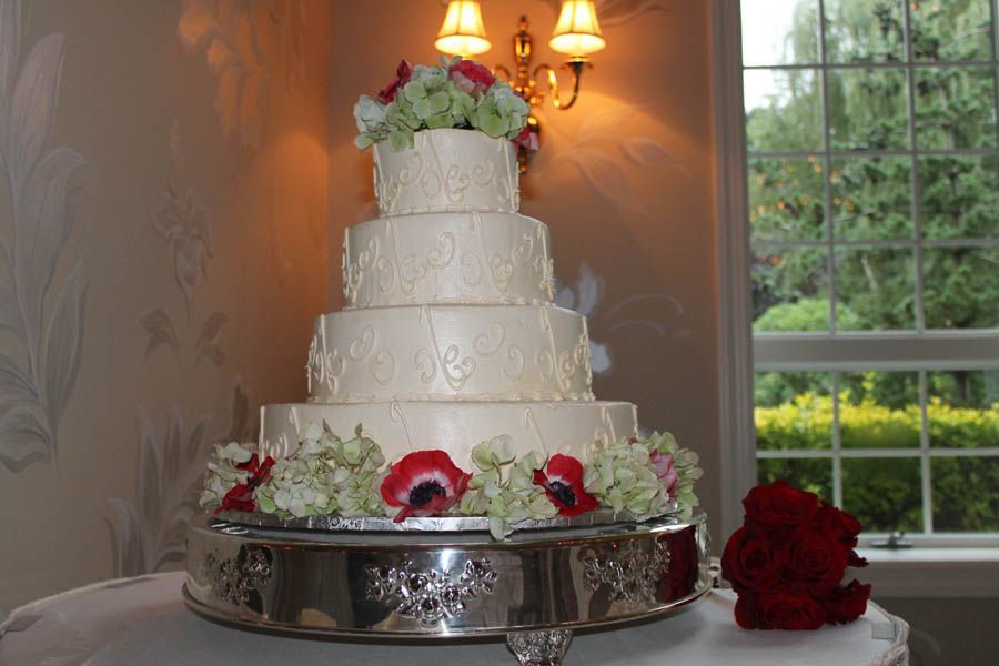 Five-tiered wedding cake with white frosting and floral decorations, on a silver stand, near a window.